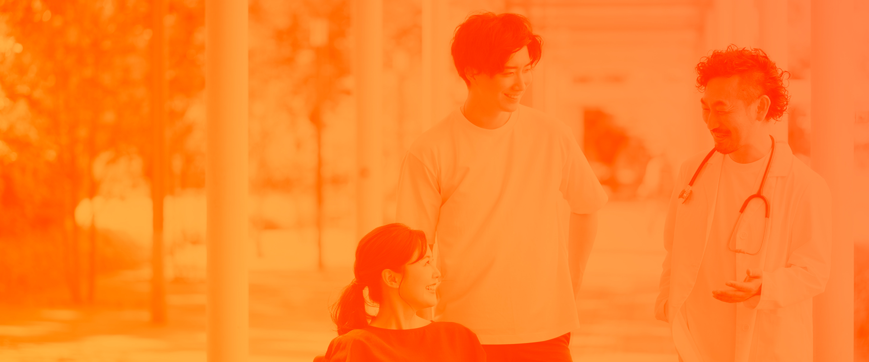 車椅子に座る女性と、その隣に立つ男性、白衣を着た医師が会話をしている